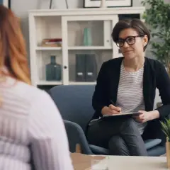 Psychoterapeutka w okularach rozmawia z pacjentką, notując coś na kartce.