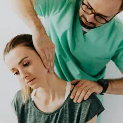 Fizjoterapeuta w zielonym fartuchu wykonuje masaż szyi młodej kobiety.