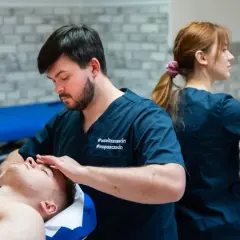 Fizjoterapeuta wykonuje masaż twarzy pacjentowi, w tle druga fizjoterapeutka.