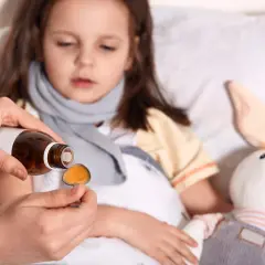 Dziecko w łóżku z gorączką, ręka podaje lek na łyżeczce.