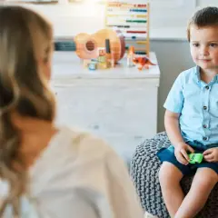 Chłopiec w niebieskiej koszuli siedzi na pufie, rozmawiając z psychoterapeutką. W tle zabawki.