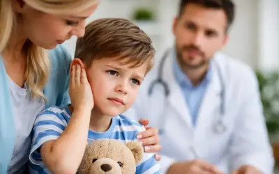 Chłopiec z pluszowym misiem trzyma się za ucho, wyraźnie cierpiąc. Mama go pociesza, a lekarz w tle bada. To zapalenie ucha u dziecka.
