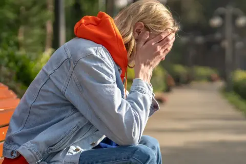 Osoba w pomarańczowej bluzie i dżinsowej kurtce siedzi na ławce, zakrywając twarz dłońmi. Może to symbolizować trudności związane z chorobą Addisona.