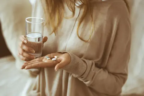 Kobieta trzyma szklankę wody i kilka tabletek, w tym naproxen Hasco, gotowa do połknięcia.