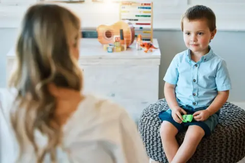 Chłopiec w niebieskiej koszuli siedzi na pufie, rozmawiając z psychoterapeutką. W tle zabawki.