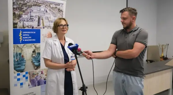Lekarka w kitlu udziela wywiadu dziennikarzowi przed banerem Uniwersyteckiego Szpitala Klinicznego w Białymstoku.