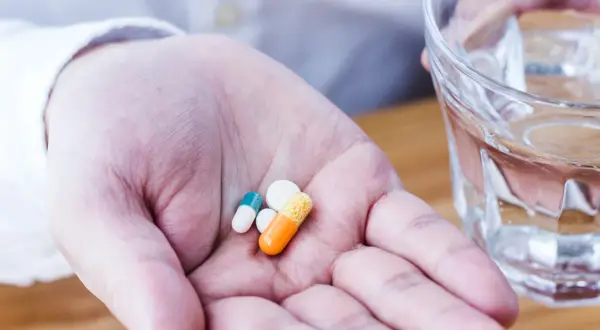 Dłoń trzymająca kilka tabletek i kapsułek obok szklanki wody.