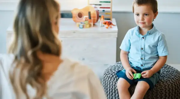 Chłopiec w niebieskiej koszuli siedzi na pufie, rozmawiając z psychoterapeutką. W tle zabawki.