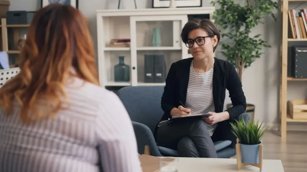 Psychoterapeutka w okularach rozmawia z pacjentką, notując coś na kartce.