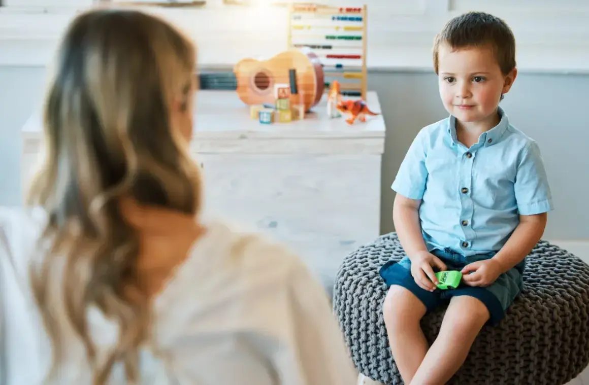 Chłopiec w niebieskiej koszuli siedzi na pufie, rozmawiając z psychoterapeutką. W tle zabawki.