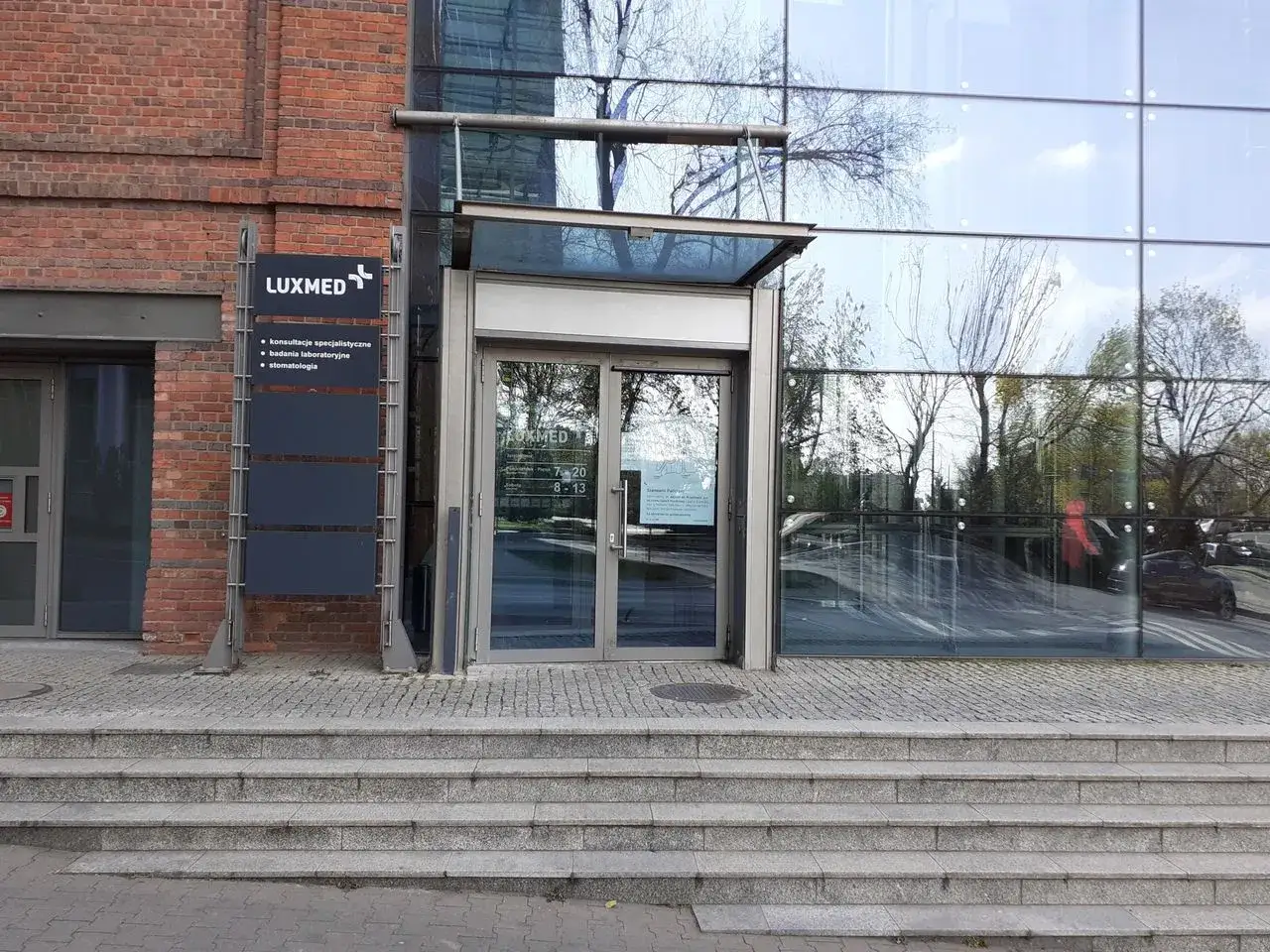 Wejście do nowoczesnego centrum medycznego Luxmed z ceglaną i szklaną fasadą.