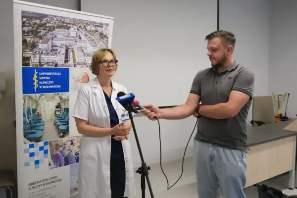 Lekarka w kitlu udziela wywiadu dziennikarzowi przed banerem Uniwersyteckiego Szpitala Klinicznego w Białymstoku.