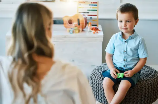 Chłopiec w niebieskiej koszuli siedzi na pufie, rozmawiając z psychoterapeutką. W tle zabawki.